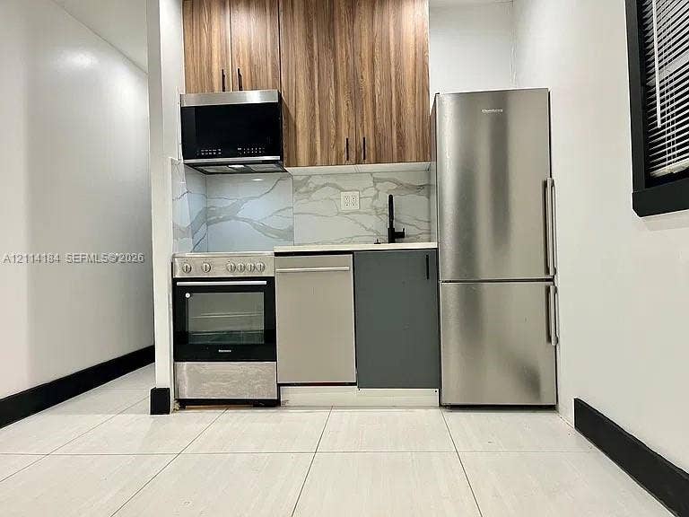 1614 Harrison Street, #1B, Hollywood, Hollywood, FL 33020 Kitchen featuring wood-finish upper cabinetry, stone-patterned backsplash, stainless steel appliances, matte black hardware, and large-format tile flooring