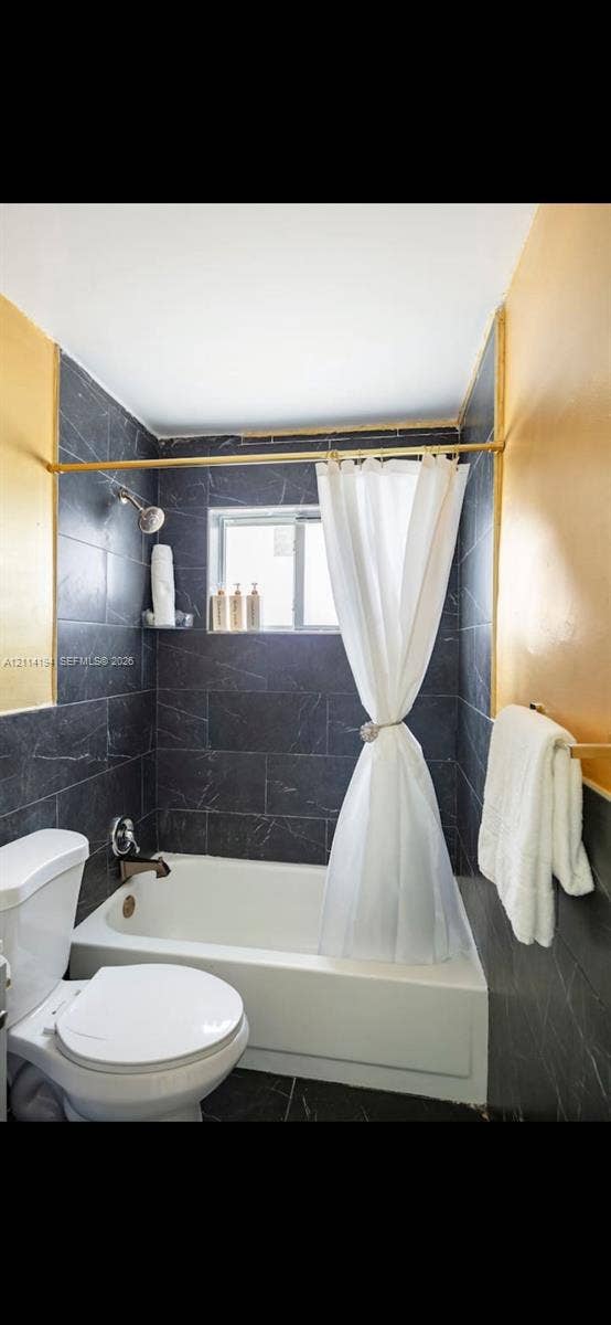 Bathroom featuring dark charcoal stone-finish wall tiling, white porcelain bathtub, metallic finish shower fixtures, and brushed-gold tone hardware
