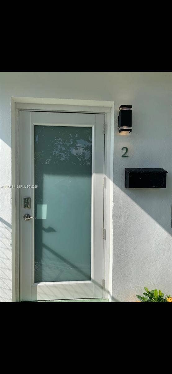 Entry door featuring a white frame, large vertical frosted glass panel, electronic deadbolt, exterior wall-mounted black lantern-style sconce, and attached black mailbox