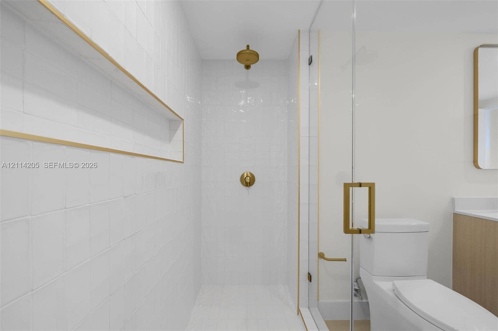 Walk-in shower with floor-to-ceiling white tile, recessed shelving with gold trim, brass fixtures, frameless glass door, and a two-piece toilet