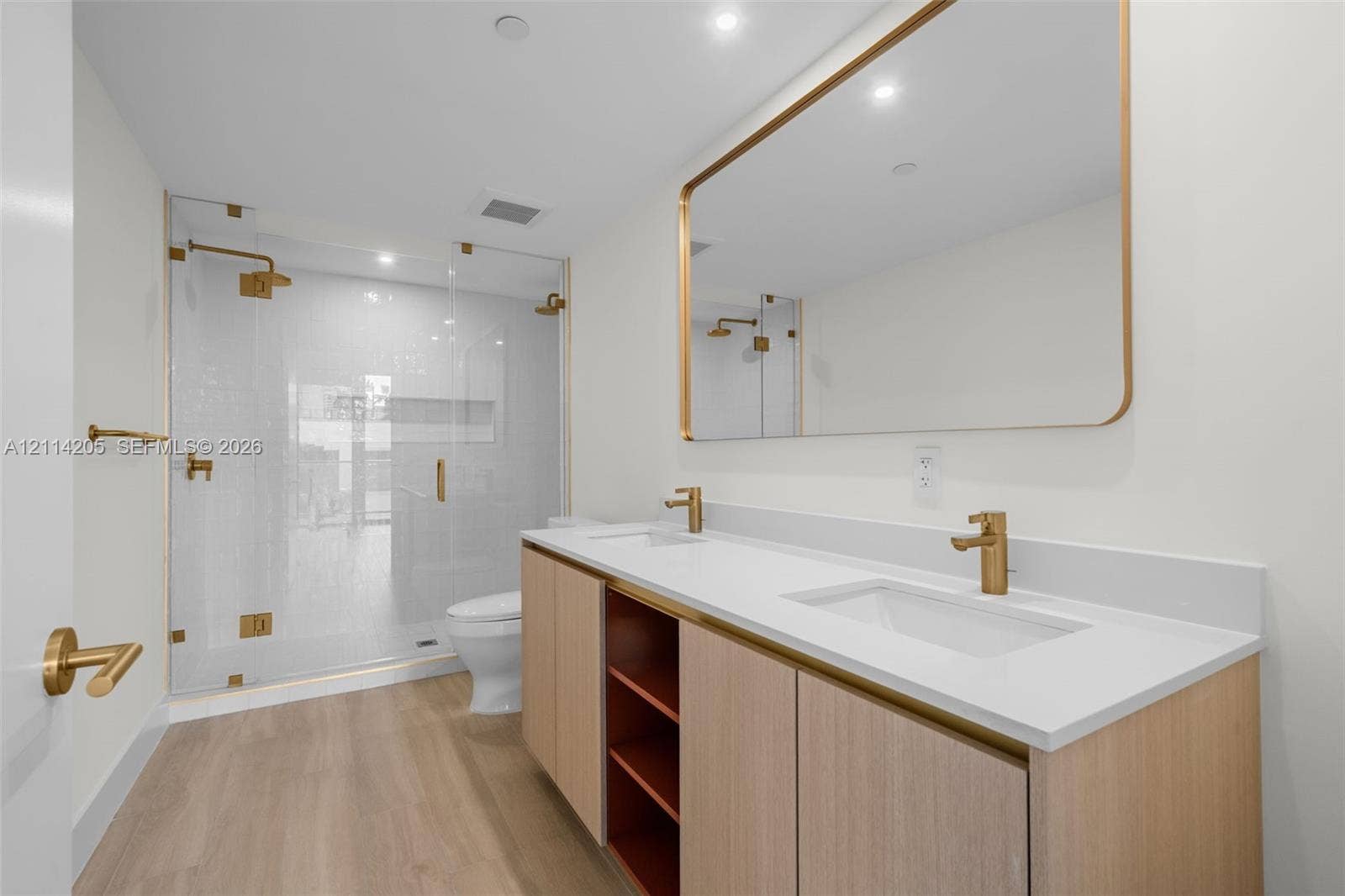 Master bathroom Dual-sink vanity with light wood cabinetry, white countertop, and gold-finish fixtures, complemented by a glass-enclosed walk-in shower with gold-finish hardware and wood-finish flooring