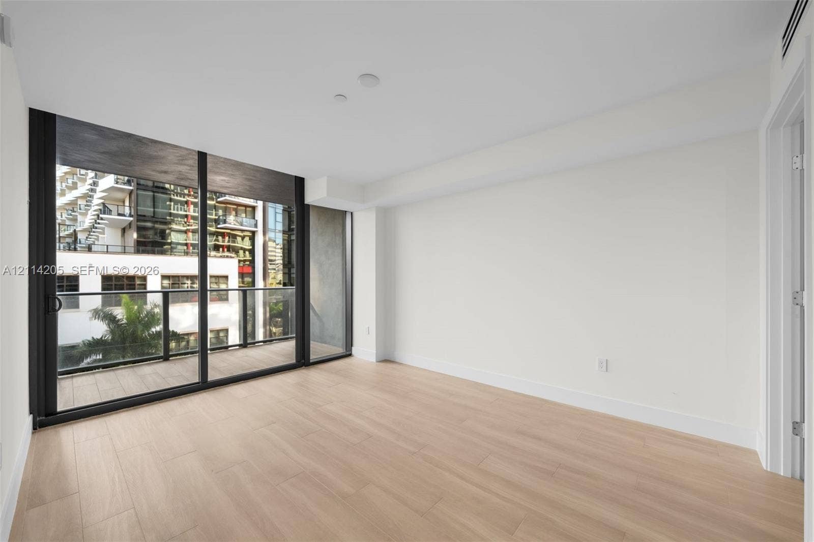 Floor-to-ceiling sliding glass doors, neutral wood-finish flooring, and an attached balcony with black metal railings