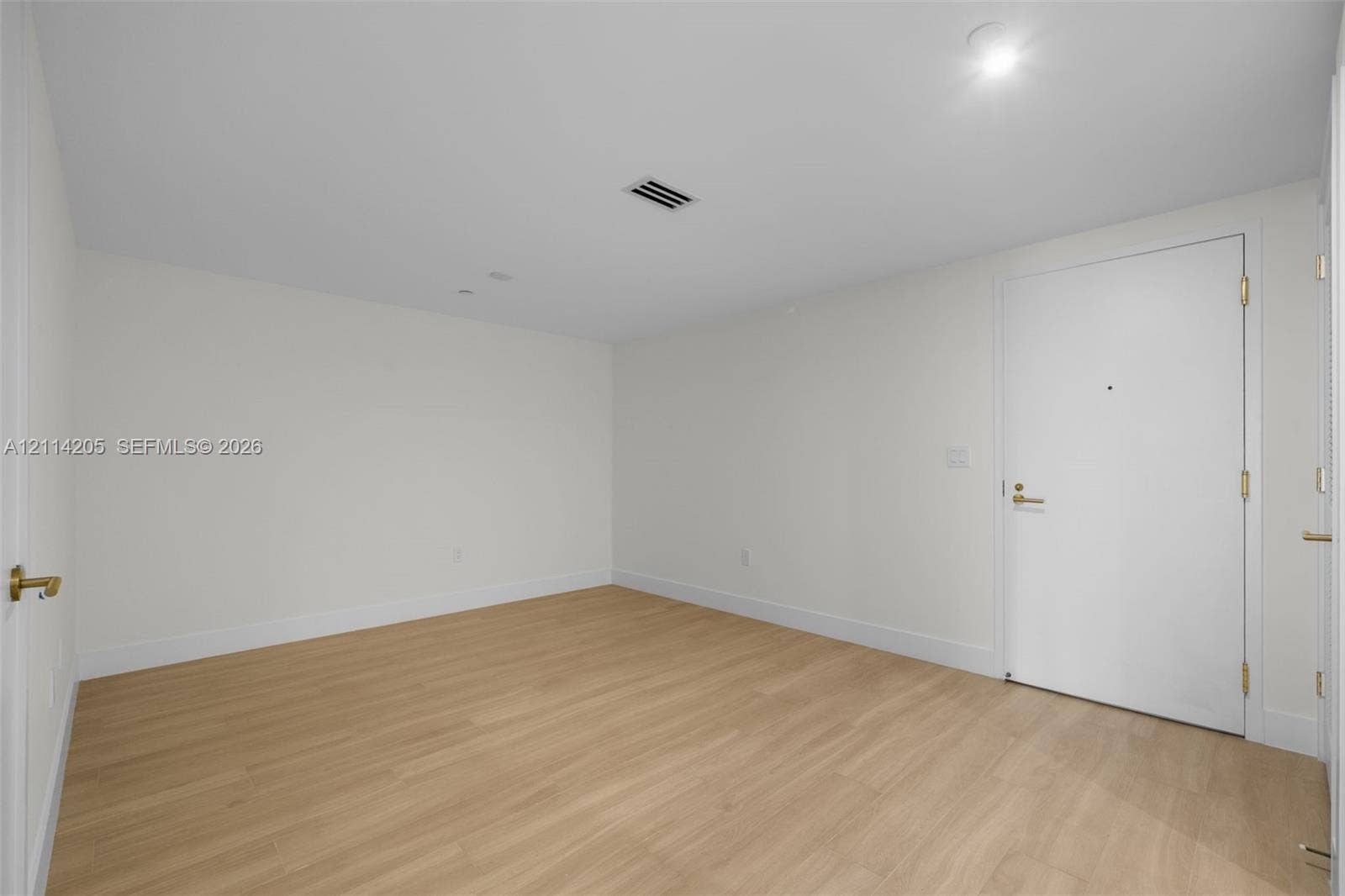Neutral-toned room featuring light wood-finish flooring, white walls, and a white interior door with metallic hardware