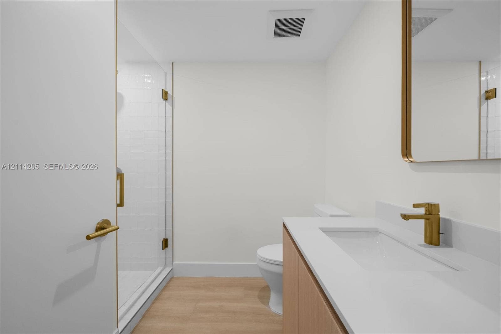Guest bathroom featuring a walk-in shower with a glass enclosure, wood-finish flooring, vanity with a white countertop and integrated sink, brass-toned hardware, and a matching framed mirror