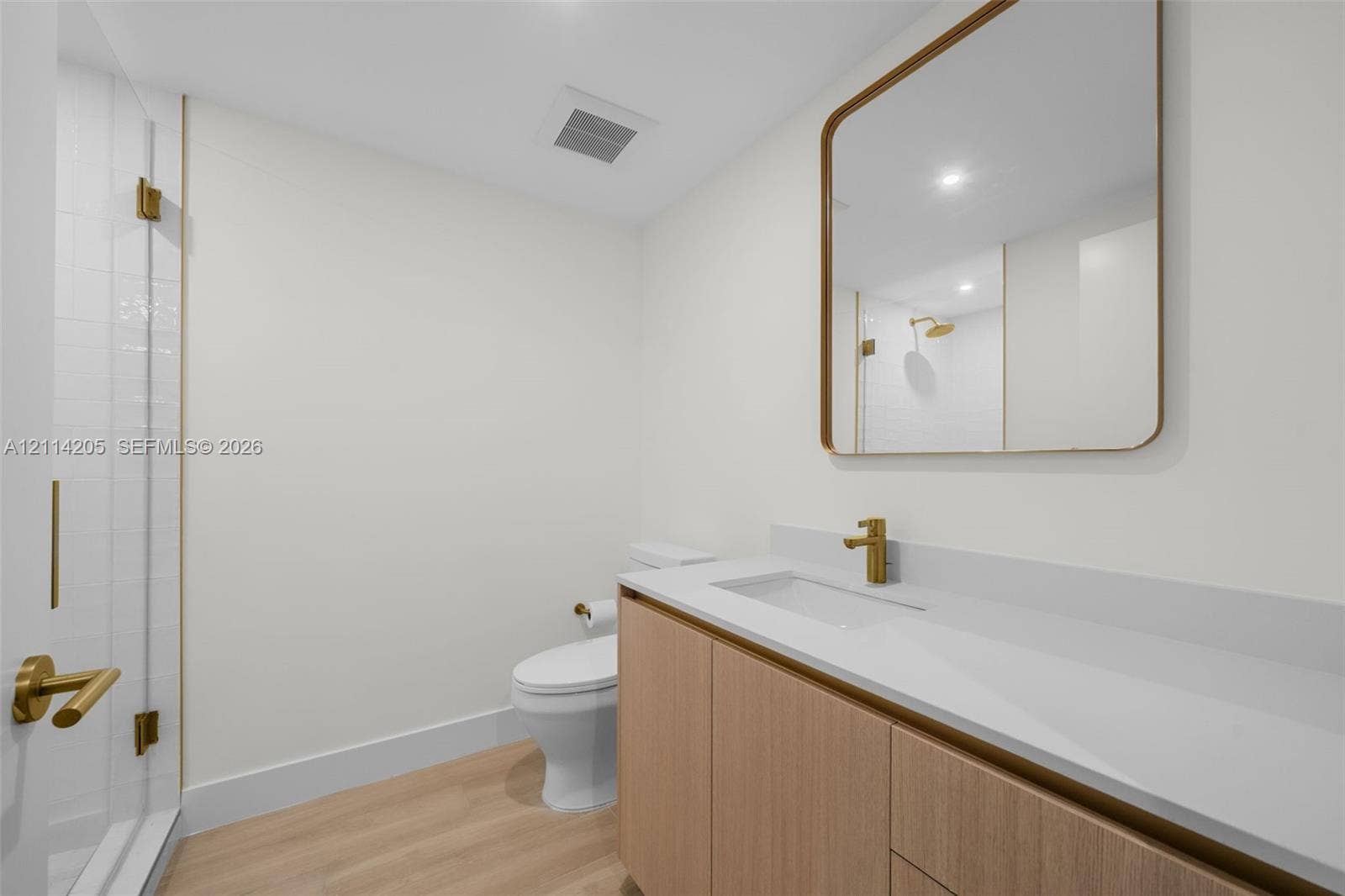Guest full bathroom featuring a wood-finish vanity, solid surface countertop, brass-toned faucet and hardware, glass-enclosed shower with tiled surround, and a rectangular mirror with a matching metallic frame