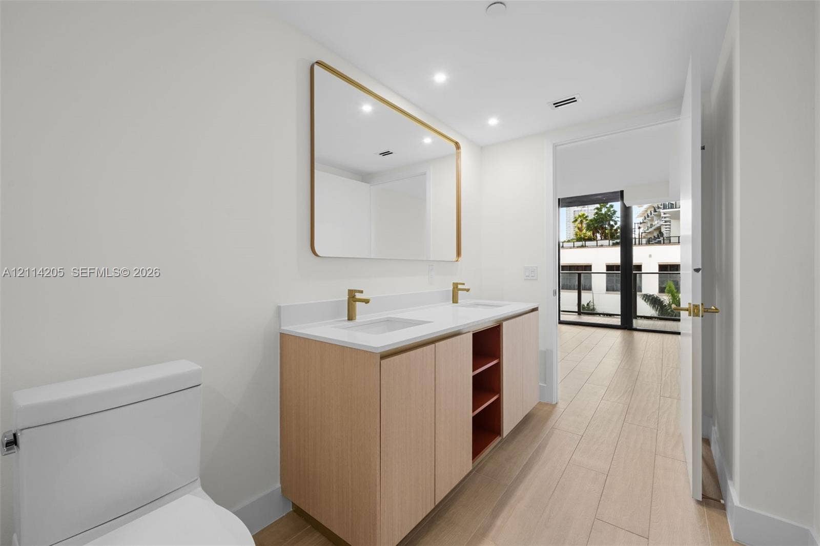 Master bathroom Dual-sink vanity with light wood cabinetry, white stone countertop, rectangular undermount sinks, and gold-tone fixtures, complemented by a large gold-framed wall mirror and wood-finish tile flooring