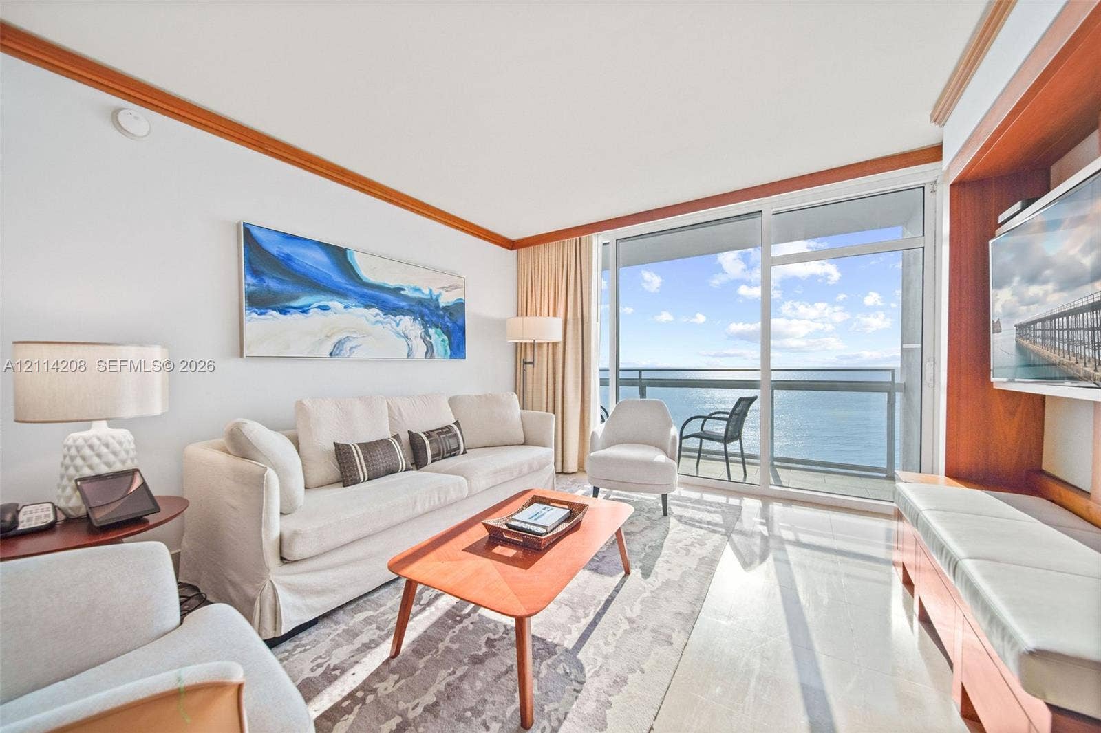 6801 Collins Avenue, #604, Miami Beach, FL 33141 Floor-to-ceiling glass sliding doors opening to an exterior balcony, built-in wooden media cabinetry, polished stone flooring, and crown molding