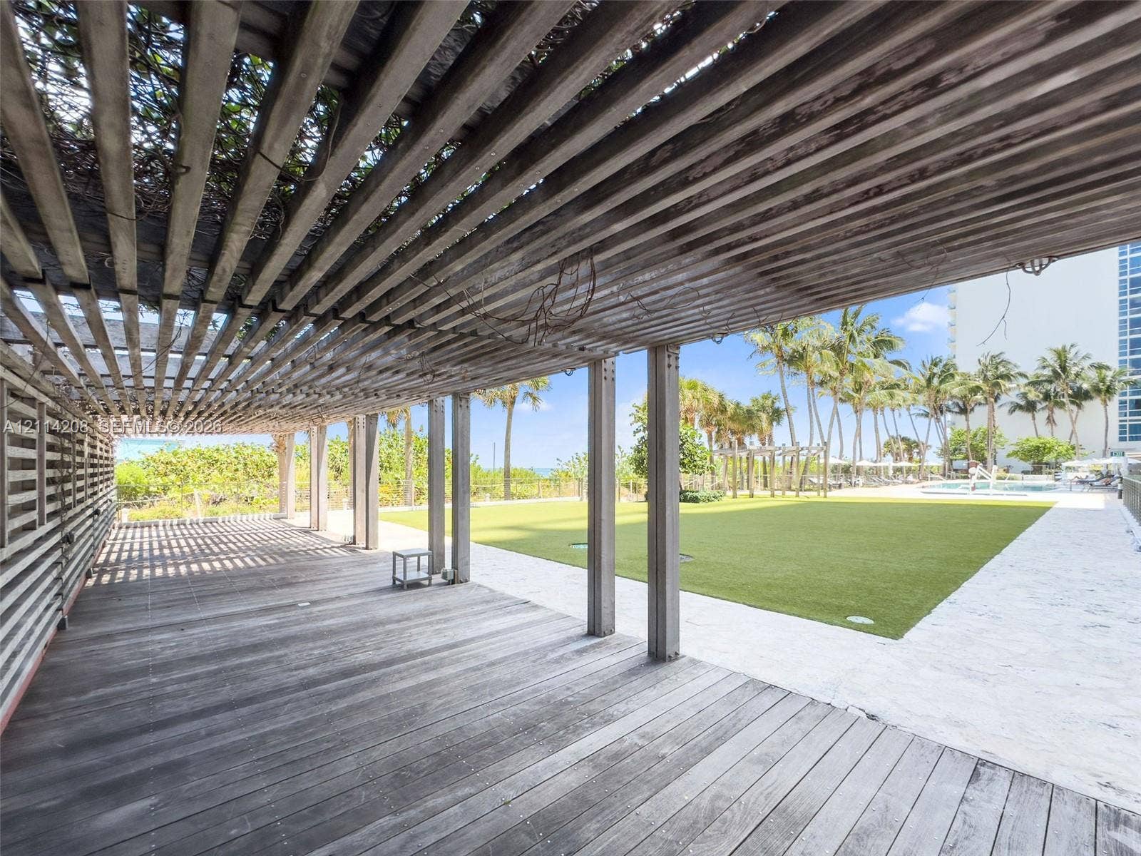 6801 Collins Avenue, #604, Miami Beach, FL 33141 Covered wood-plank patio featuring a slatted pergola structure, synthetic turf lawn, stone-paver walkways, and mature palm trees