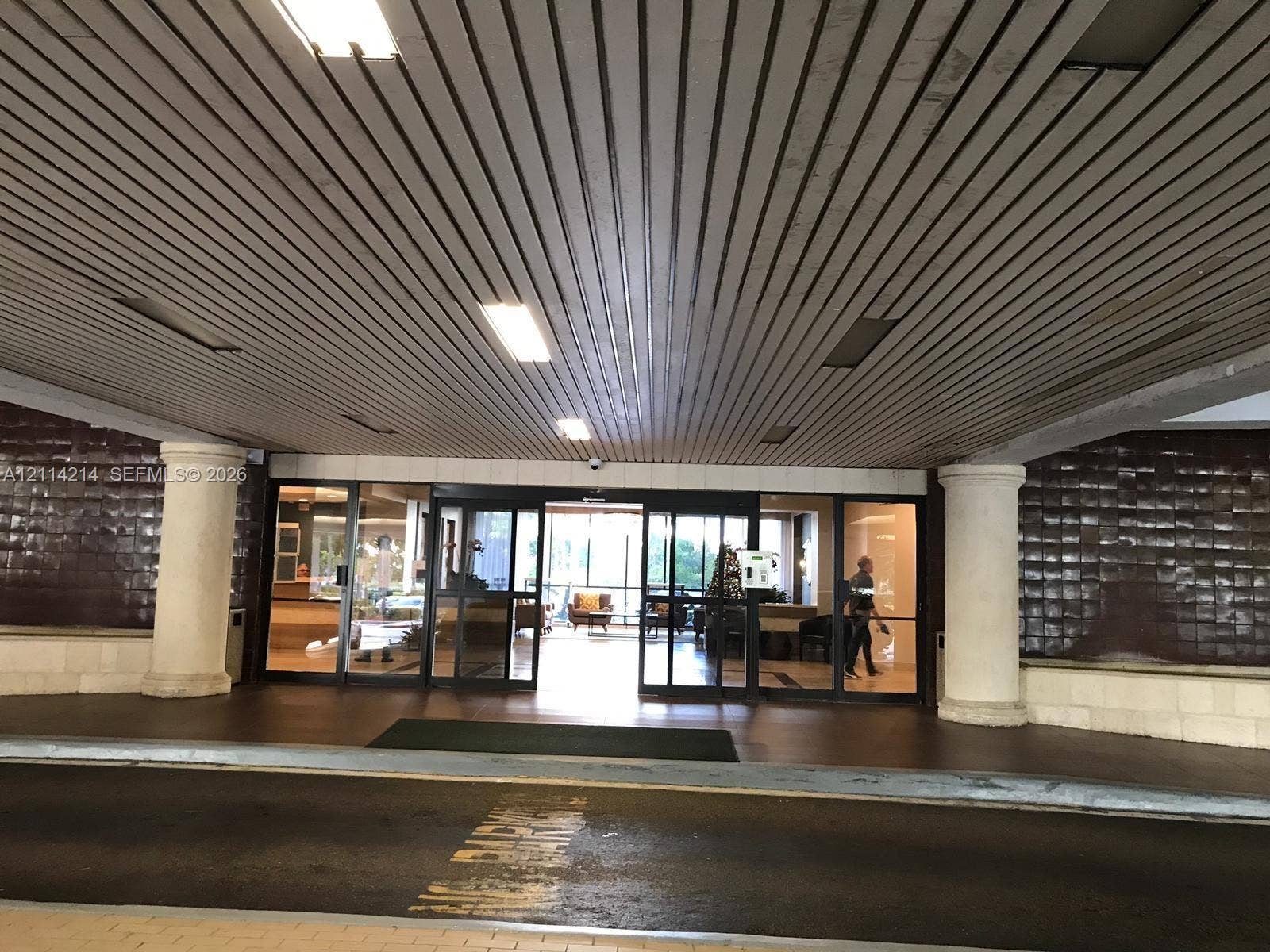 3701 N Country Club Drive, #706, Aventura, FL 33180 Covered drive-up entrance featuring a slatted ceiling, prominent architectural columns, and expansive glass-paneled sliding entry doors with dark-toned tile exterior siding
