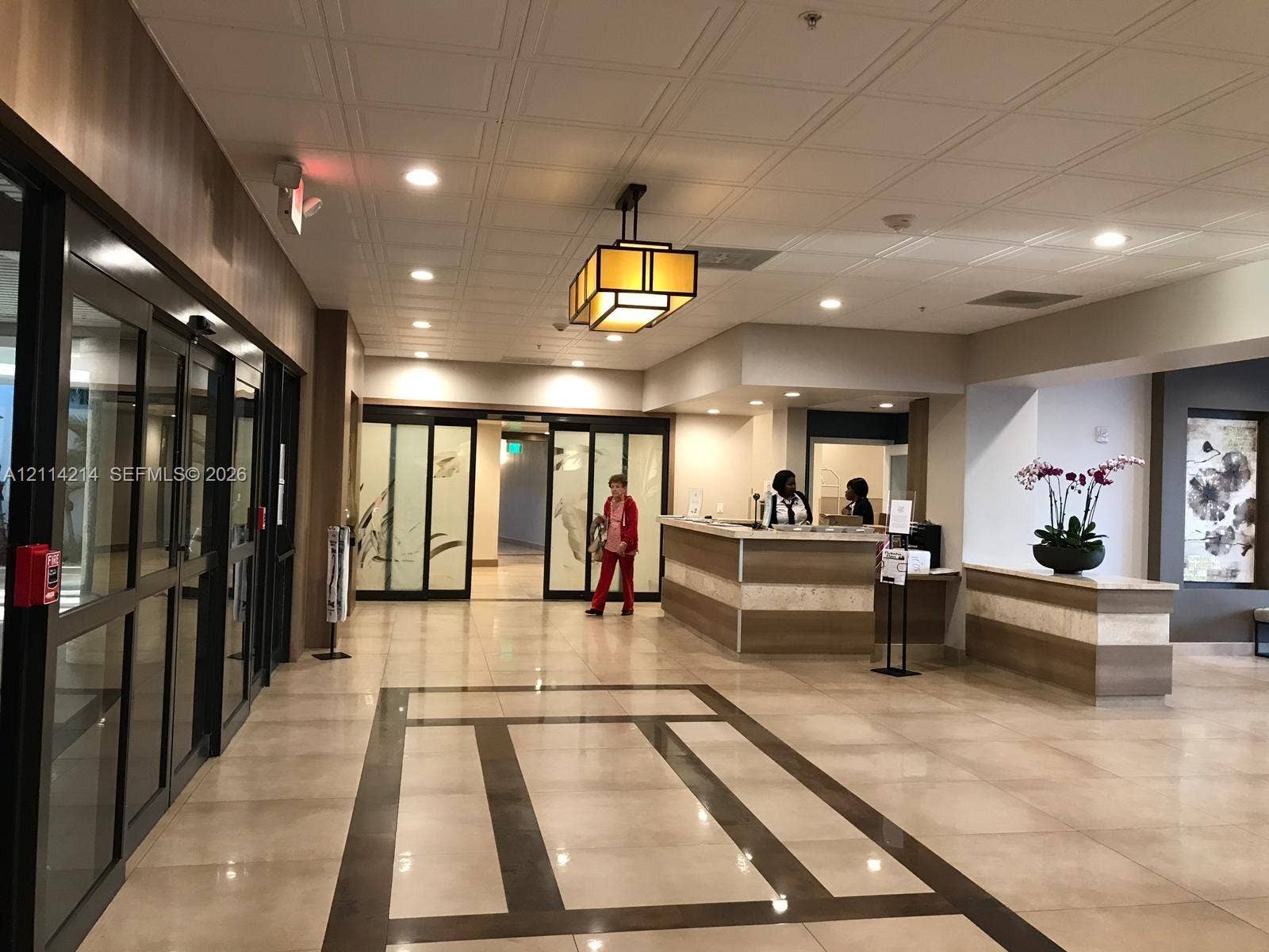 3701 N Country Club Drive, #706, Aventura, FL 33180 Lobby featuring tile flooring with decorative stone inlay, coffered ceiling, custom reception desk with stone detailing, glass-paneled sliding entry doors, and geometric mission-style pendant lighting