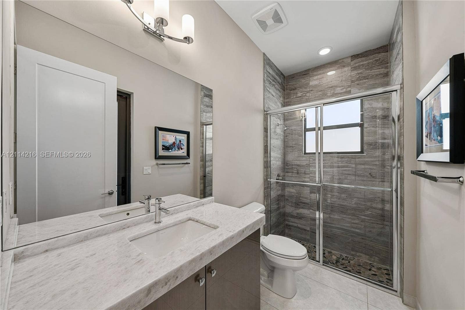 17085 Teton River Road, Boca Raton, FL 33496 Vanity with light-toned stone-finish countertop and integrated rectangular basin, full-width mirror, glass-enclosed shower with horizontal textured tile surround, and chrome hardware