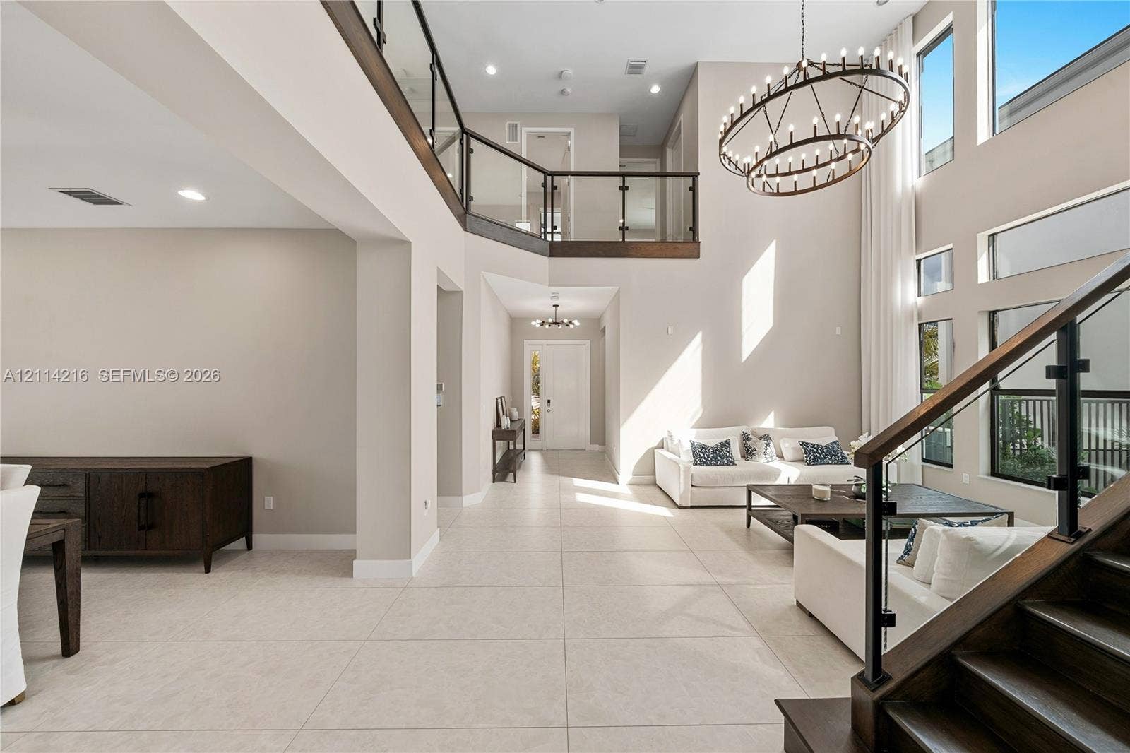 17085 Teton River Road, Boca Raton, FL 33496 Double-height ceiling, open-concept floor plan with large-format tile flooring, staircase with dark wood treads and glass railings, multi-tiered circular chandelier, and expansive vertical windows