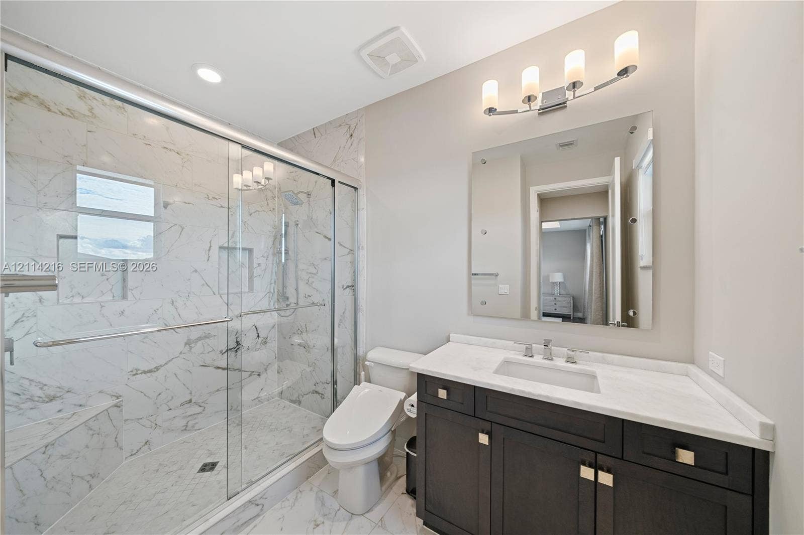 17085 Teton River Road, Boca Raton, FL 33496 Bathroom featuring a glass-enclosed walk-in shower with stone-patterned tile, dark wood-finish cabinetry, a white countertop with an integrated rectangular sink, and a wall-mounted vanity mirror with a four-light fixture