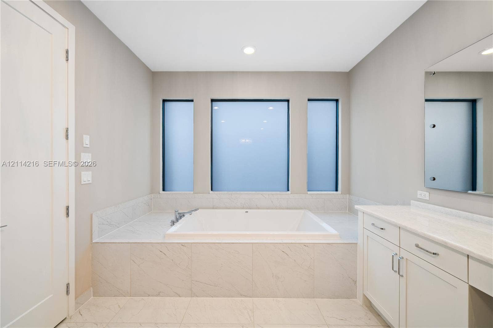 17085 Teton River Road, Boca Raton, FL 33496 Built-in soaking tub with a tiled surround, triple-window configuration, white cabinetry with stone-finish countertops, and wall-mounted vanity mirror