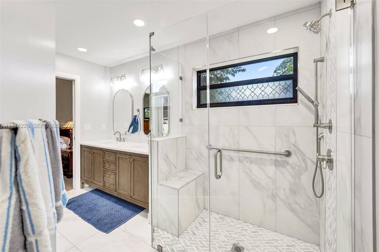 5741 SW 88th Terrace, Cooper City, FL 33328 Bathroom featuring a dual-sink vanity, glass-enclosed shower with a tiled bench, herringbone-patterned shower flooring, and wall-mounted plumbing fixtures