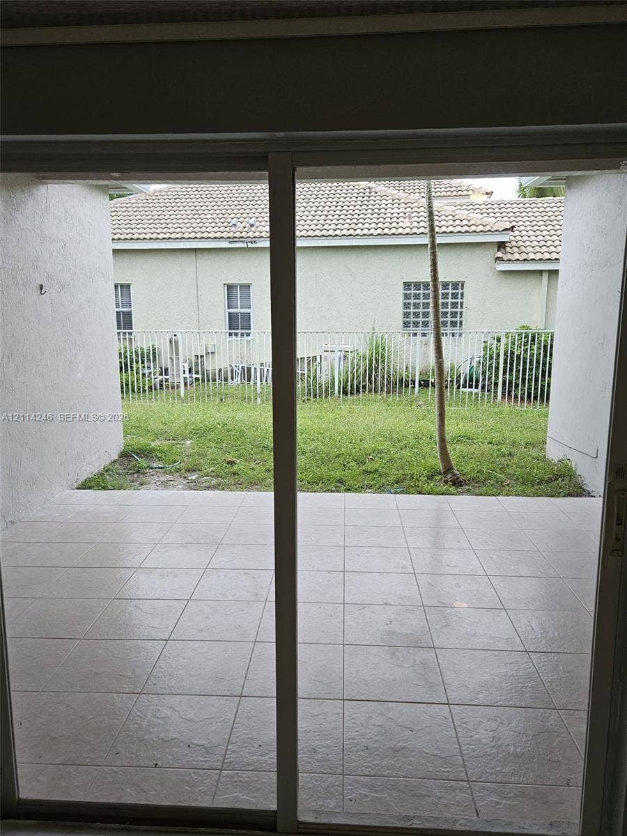 Covered patio with neutral-toned square tile flooring, enclosed exterior space with a white metal fence, and grass lawn