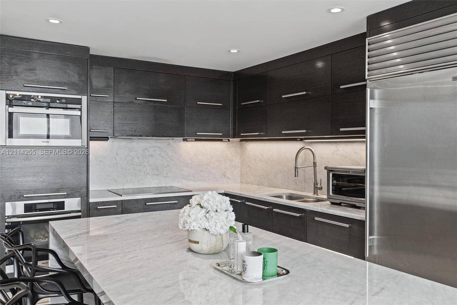 900 Biscayne Boulevard, #1705, Miami, FL 33132 Custom dark-finish cabinetry featuring horizontal bar pulls, expansive stone-surface center island, integrated stainless steel refrigeration, built-in wall oven and microwave combination, and stone-surface backsplash with under-cabinet lighting