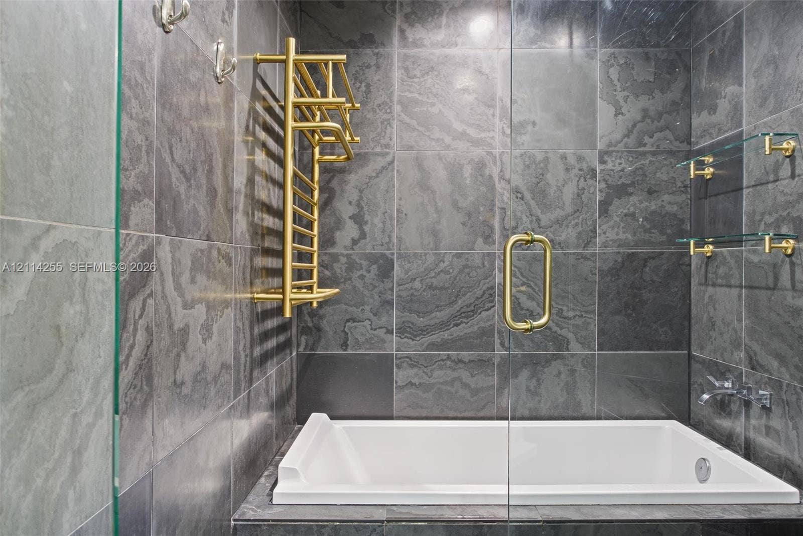 900 Biscayne Boulevard, #1705, Miami, FL 33132 Large-format charcoal stone tile surround with a white deep-soaking tub, brass-finish heated towel rack, wall-mounted faucet, and glass shelving