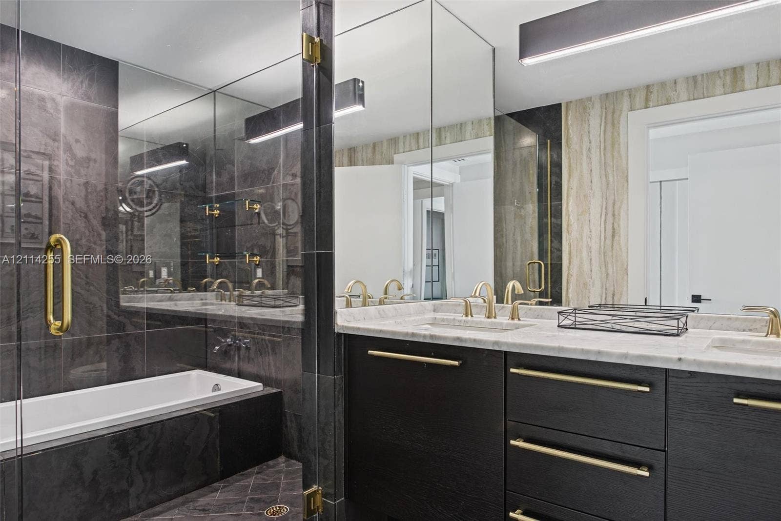 900 Biscayne Boulevard, #1705, Miami, FL 33132 Custom dark wood-finish cabinetry featuring brass hardware, dual undermount sinks, light-toned stone vanity countertop, and a walk-in shower with floor-to-ceiling dark tile surrounds and glass enclosure