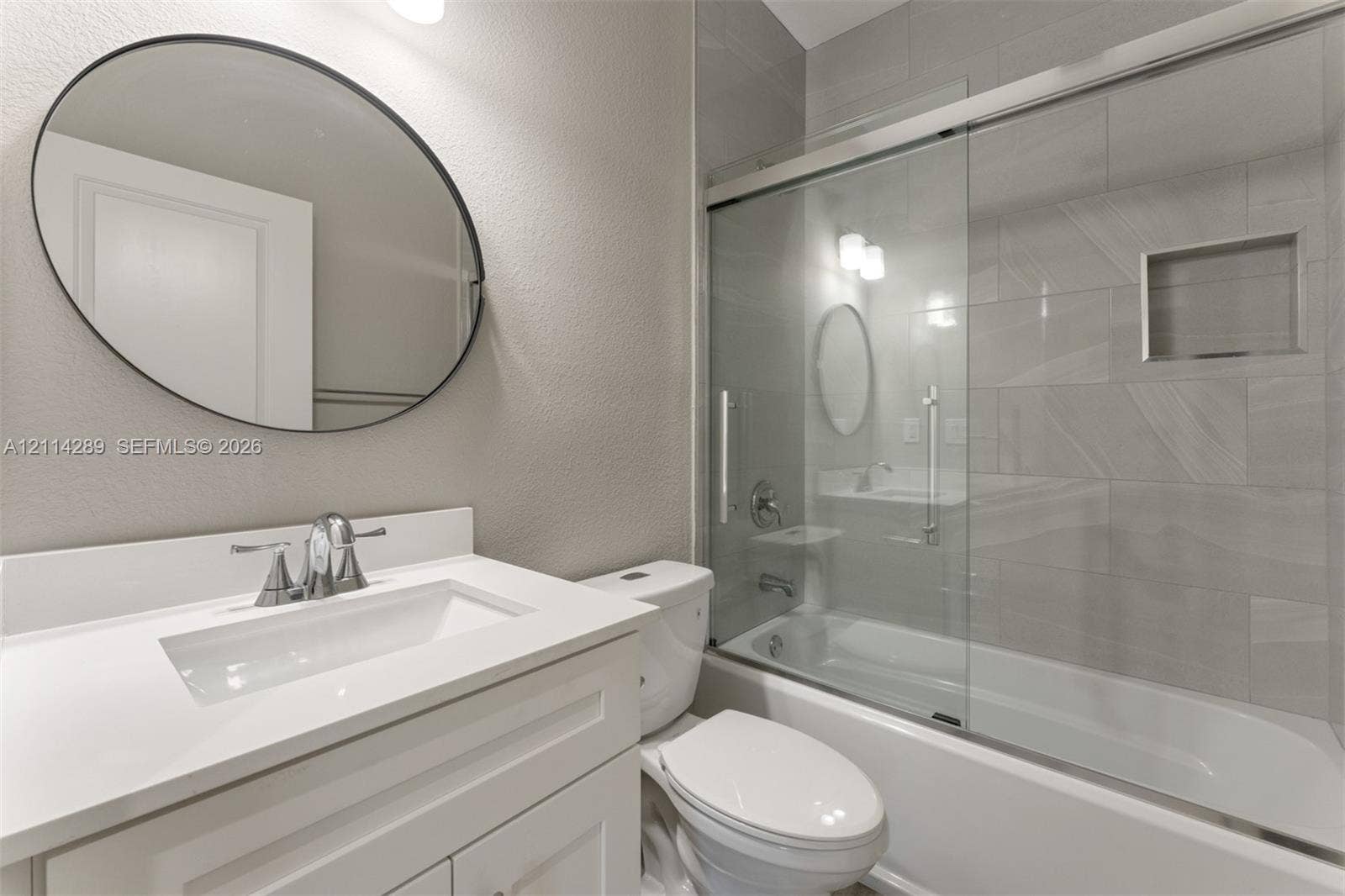 646 Cabrera Street, Key Largo, FL 33037 Bathroom featuring a vanity with a white countertop and rectangular undermount sink, oval wall mirror, sliding glass shower door, and tile-surround tub with a built-in recessed niche