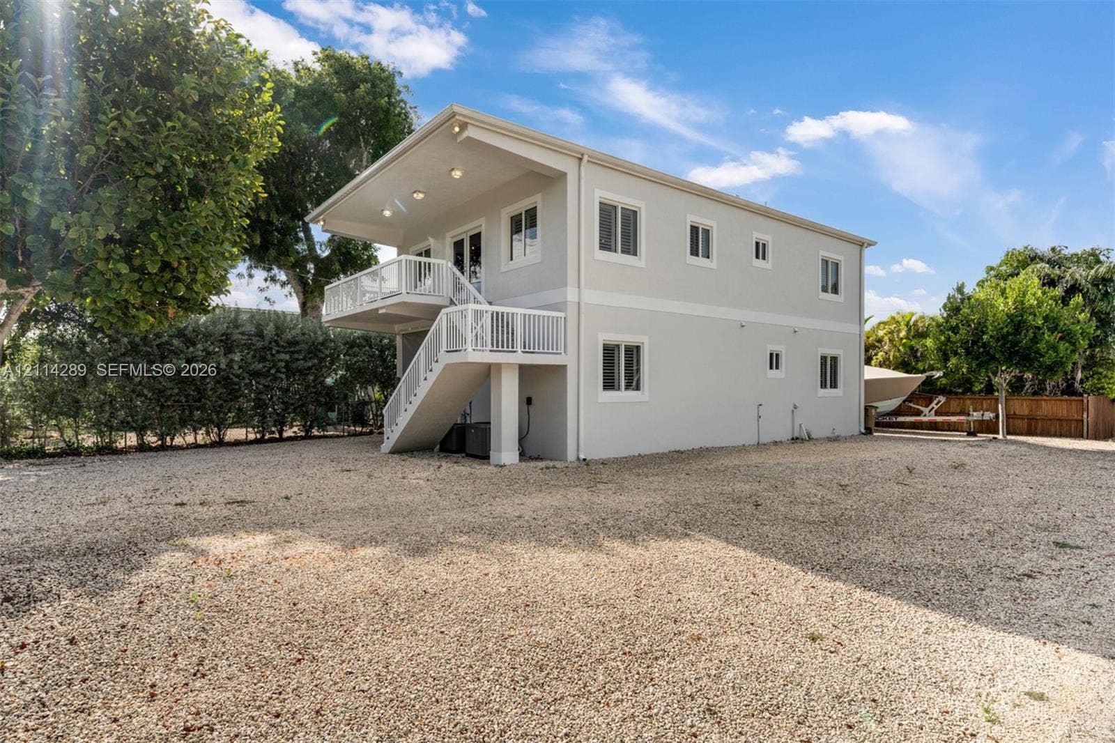646 Cabrera Street, Key Largo, FL 33037 Two-story structure featuring light-toned smooth siding, a raised upper-level deck with white railings, an exterior staircase, and a sprawling gravel grounds area