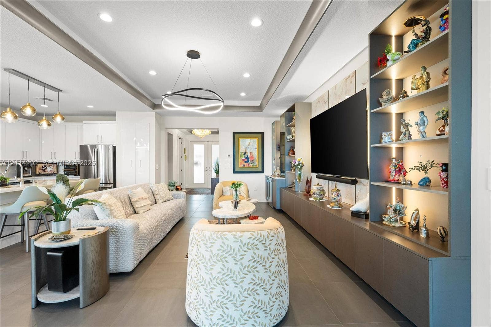 Open-concept living space featuring a recessed tray ceiling with an integrated LED chandelier, large-format tile flooring, and custom built-in cabinetry with illuminated shelving