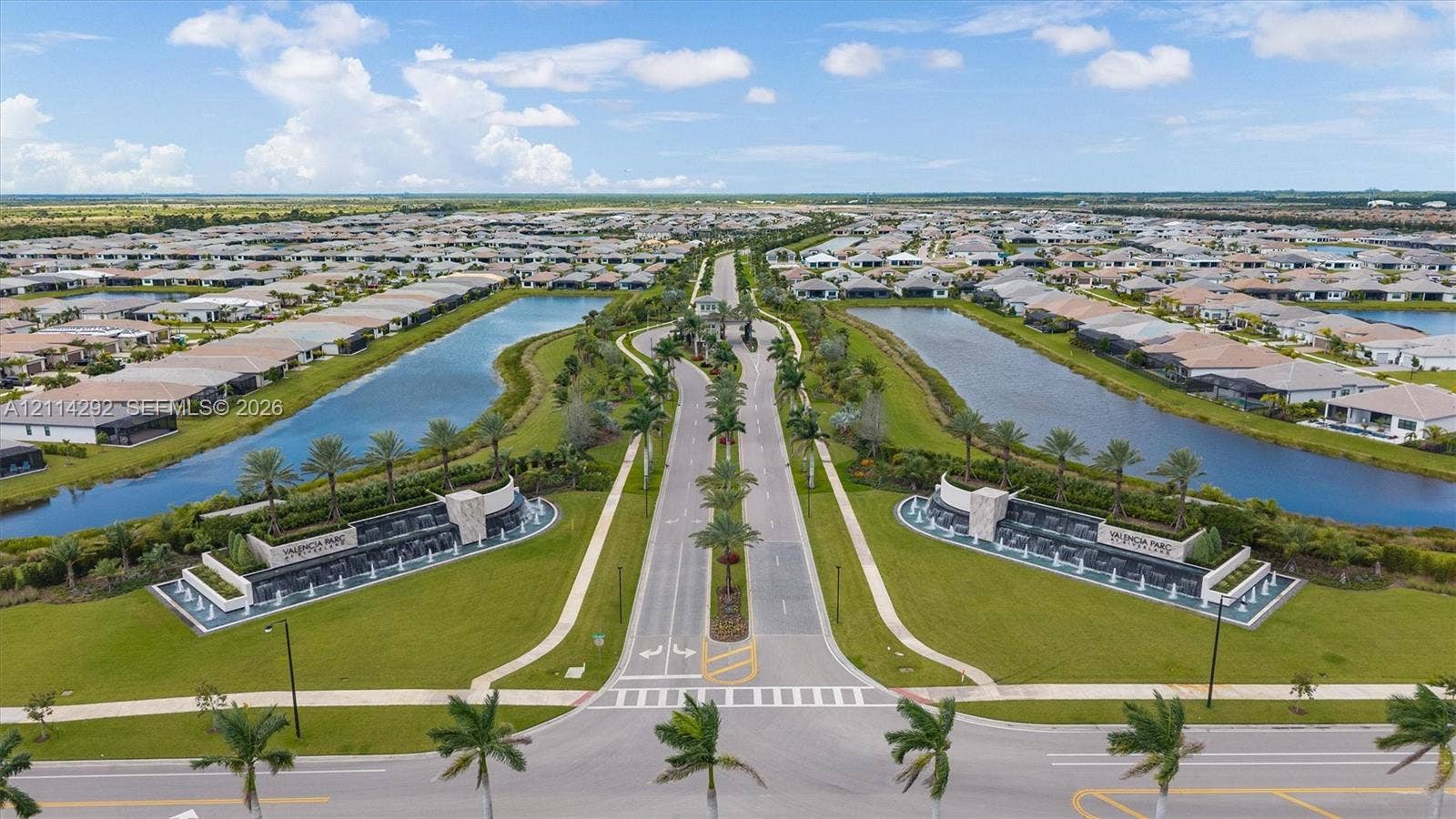Aerial perspective of community entrance featuring tiered water fountains, stone-clad monument signage, manicured landscaping, palm tree-lined boulevards, and expansive retention ponds