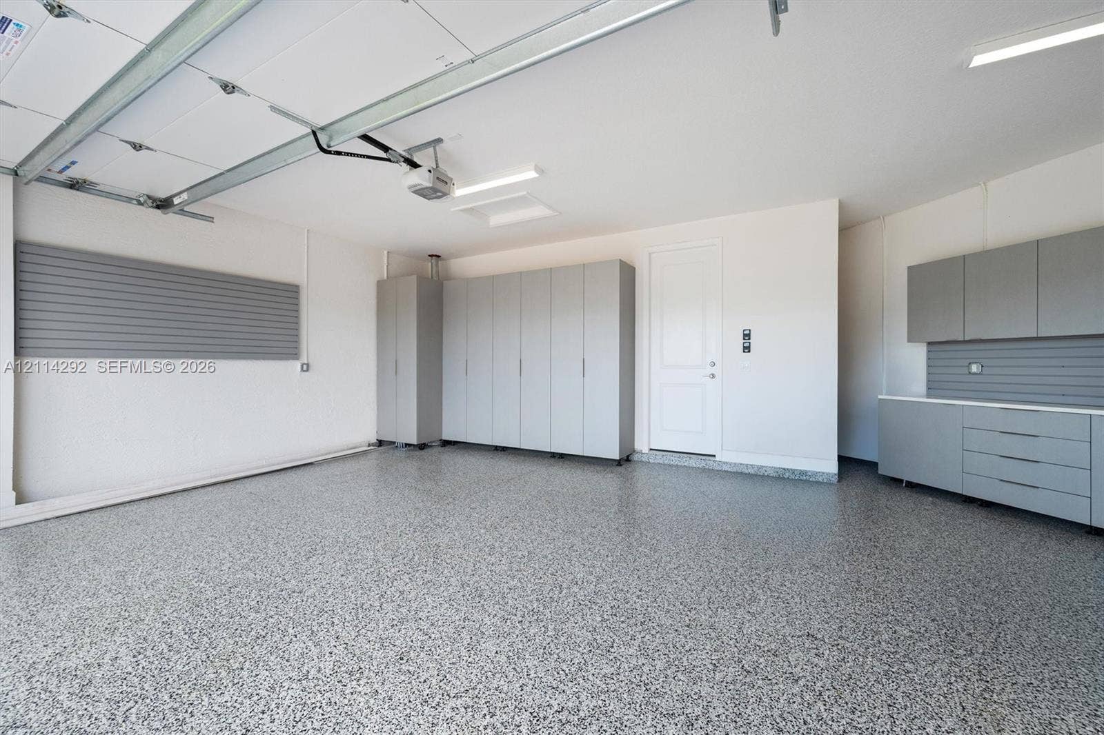 Finished garage featuring speckled epoxy flooring, built-in grey cabinetry, wall-mounted storage panels, and overhead LED lighting