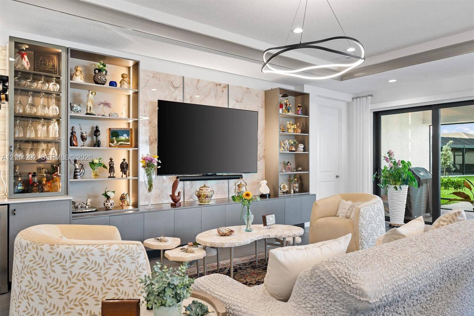 Custom built-in cabinetry featuring illuminated glass-front display shelving, stone-patterned accent wall, modern multi-ring chandelier, tray ceiling, and sliding glass patio doors