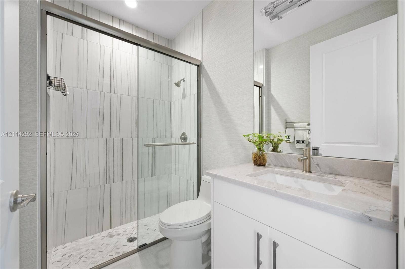 Bathroom featuring a walk-in glass shower with vertical-patterned wall tile, stone-surface countertop, integrated rectangular sink, vanity with white cabinetry, and brushed-metal hardware