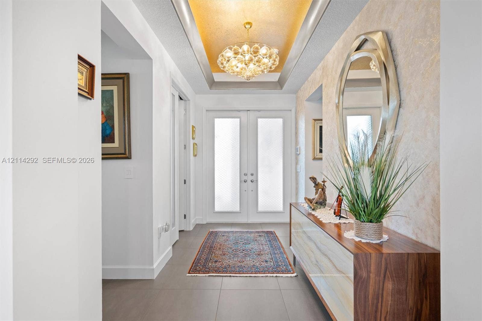 Foyer featuring double entry doors with frosted glass inserts, a recessed tray ceiling with metallic gold-tone finish, a multi-bulb tiered chandelier, and neutral large-format tile flooring
