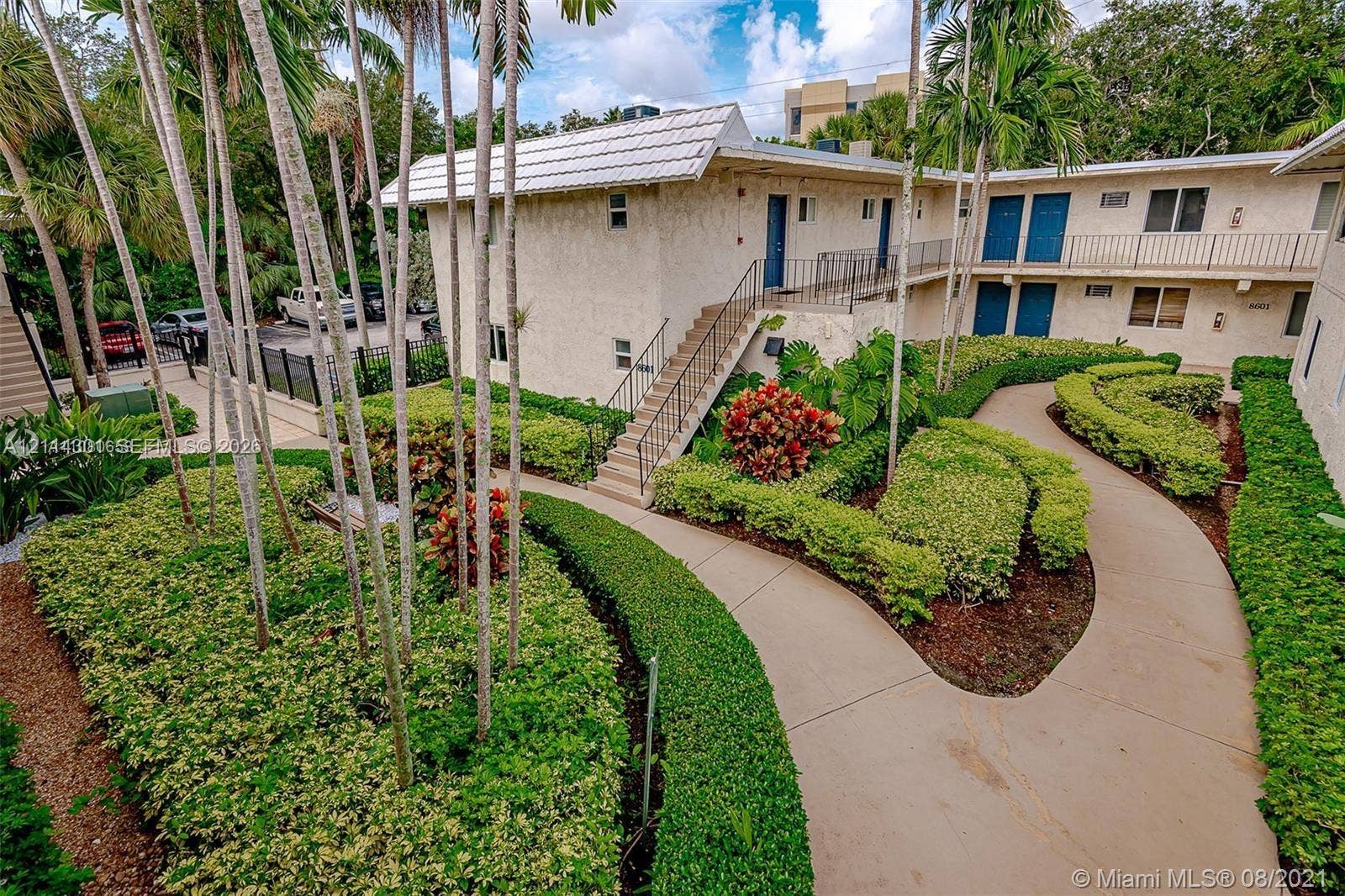 Two-story exterior featuring a light-toned stucco facade, metal staircase, expansive landscaped garden with manicured hedges, and paved walkways