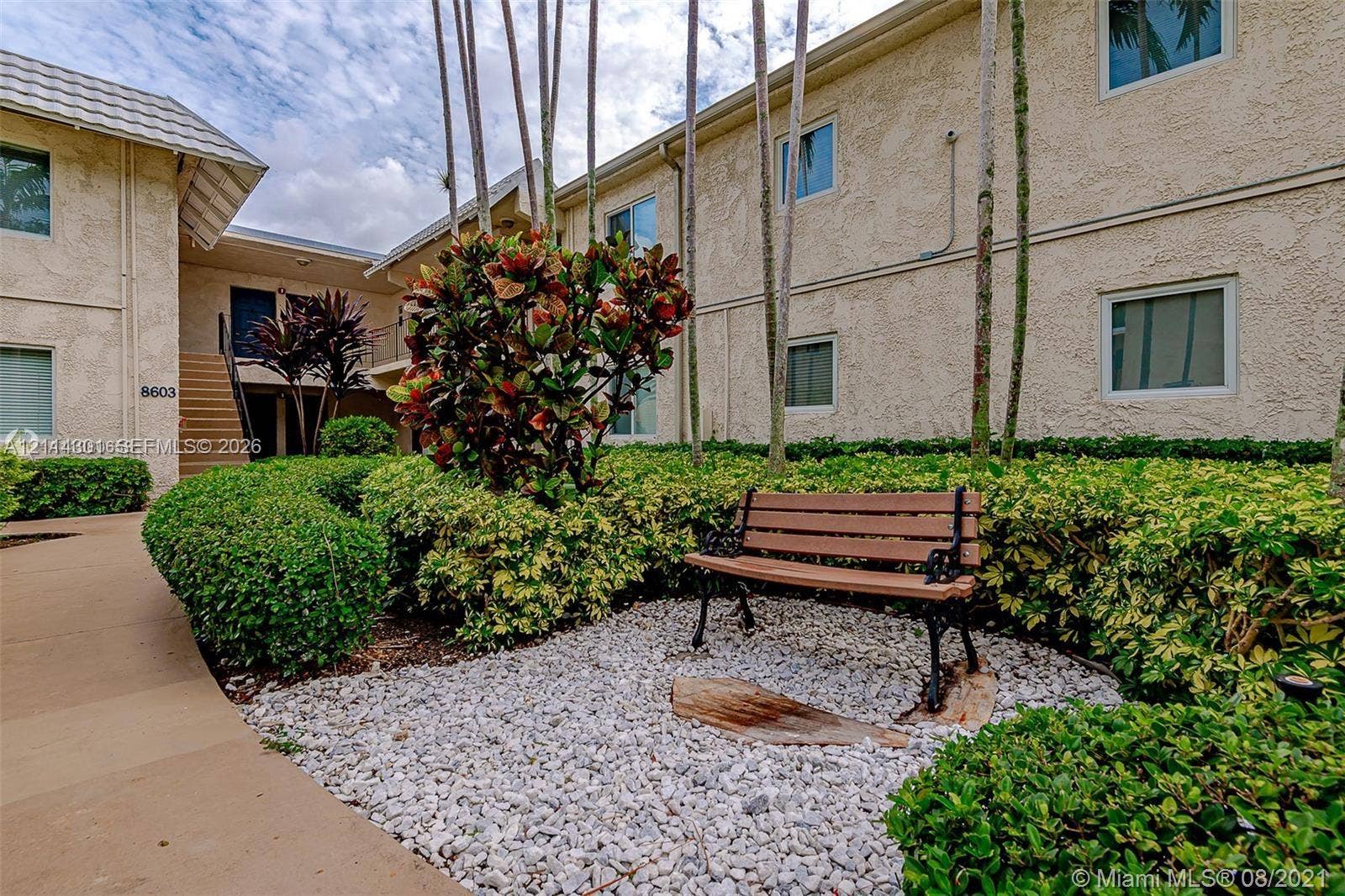 Stucco exterior building featuring a paved walkway, landscaped shrubbery, mature palms, and a rock-ground covering