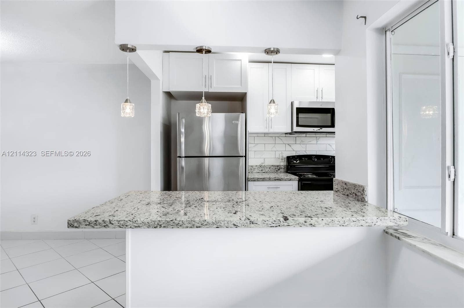 Kitchen featuring a peninsula with stone-finish countertop, three glass pendant lights, white shaker-style cabinetry, stainless steel appliances, and subway tile backsplash