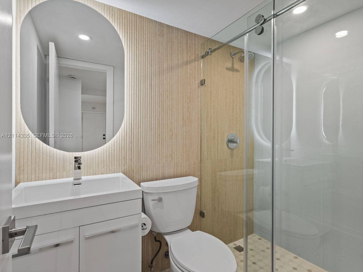 2930 Day Avenue, #N102, Miami, Miami, FL 33133 Bathroom featuring a white vanity, oval backlit mirror, vertical fluted wall paneling, and a glass-enclosed shower with mosaic floor tile