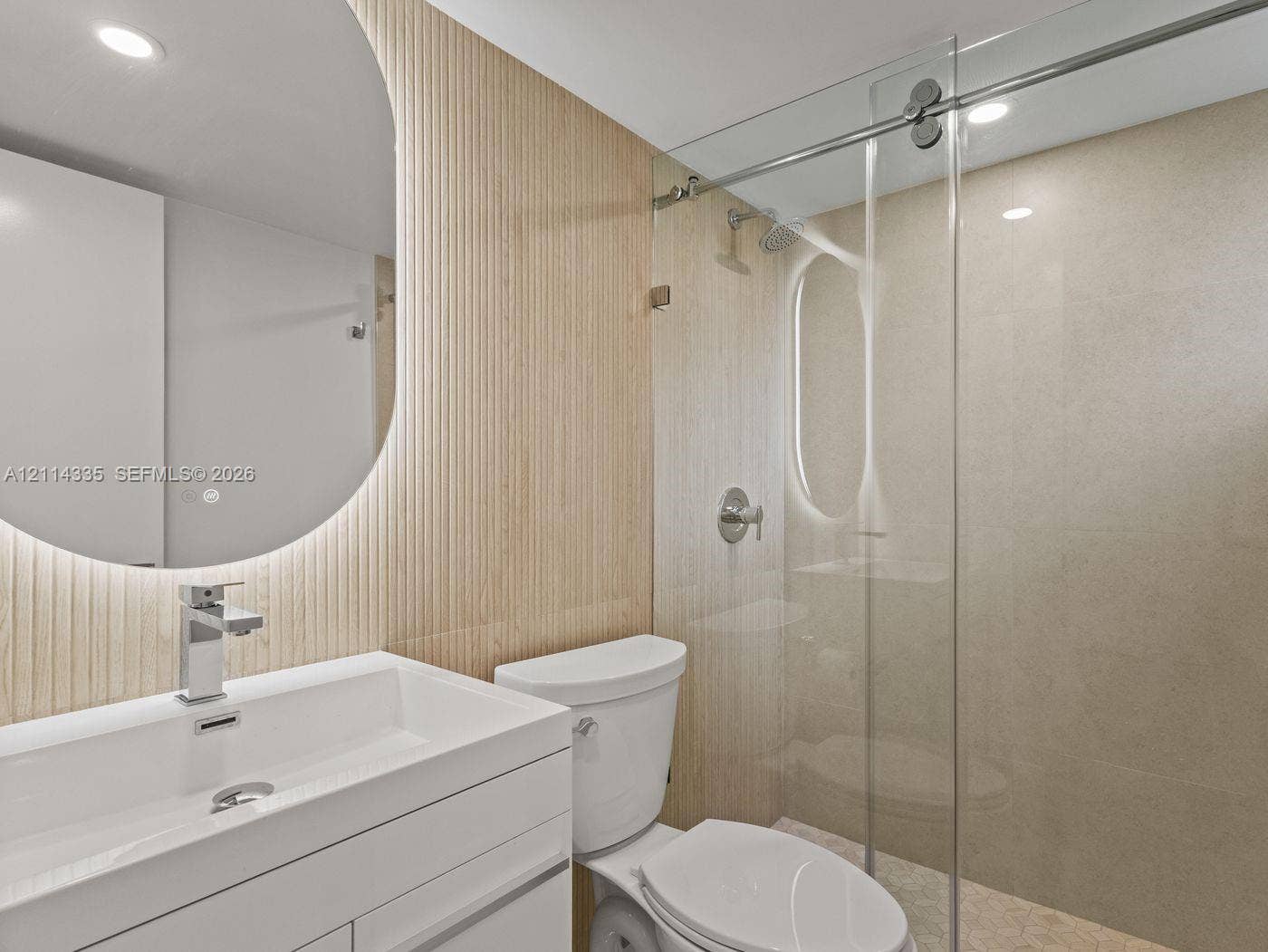 2930 Day Avenue, #N102, Miami, Miami, FL 33133 Bathroom featuring a white floating vanity, ribbed wood-panel accent wall, integrated circular mirror with backlighting, and a glass-enclosed shower with polished metal hardware