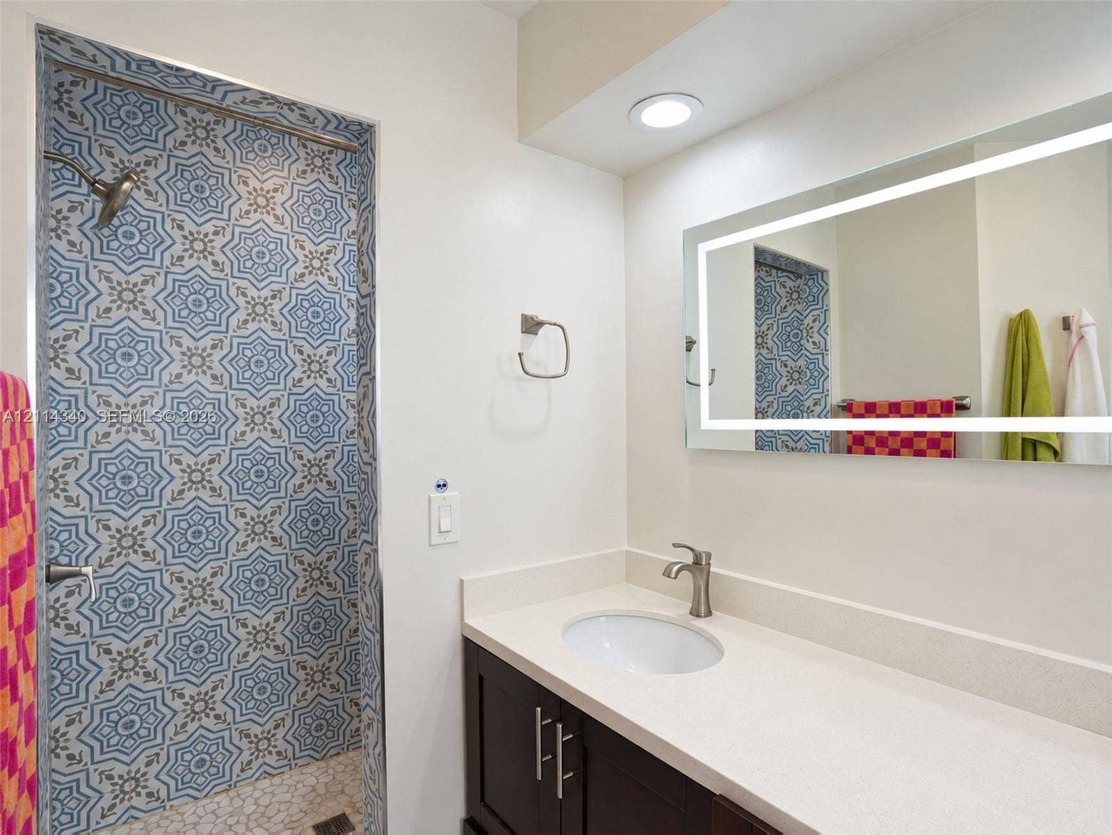 3586 SW 50th Street, Hollywood, FL 33312 Bathroom featuring a shower stall with patterned tile walls and stone-tiled flooring, dark-finish cabinetry, a solid-surface countertop with an integrated basin, and an illuminated rectangular mirror