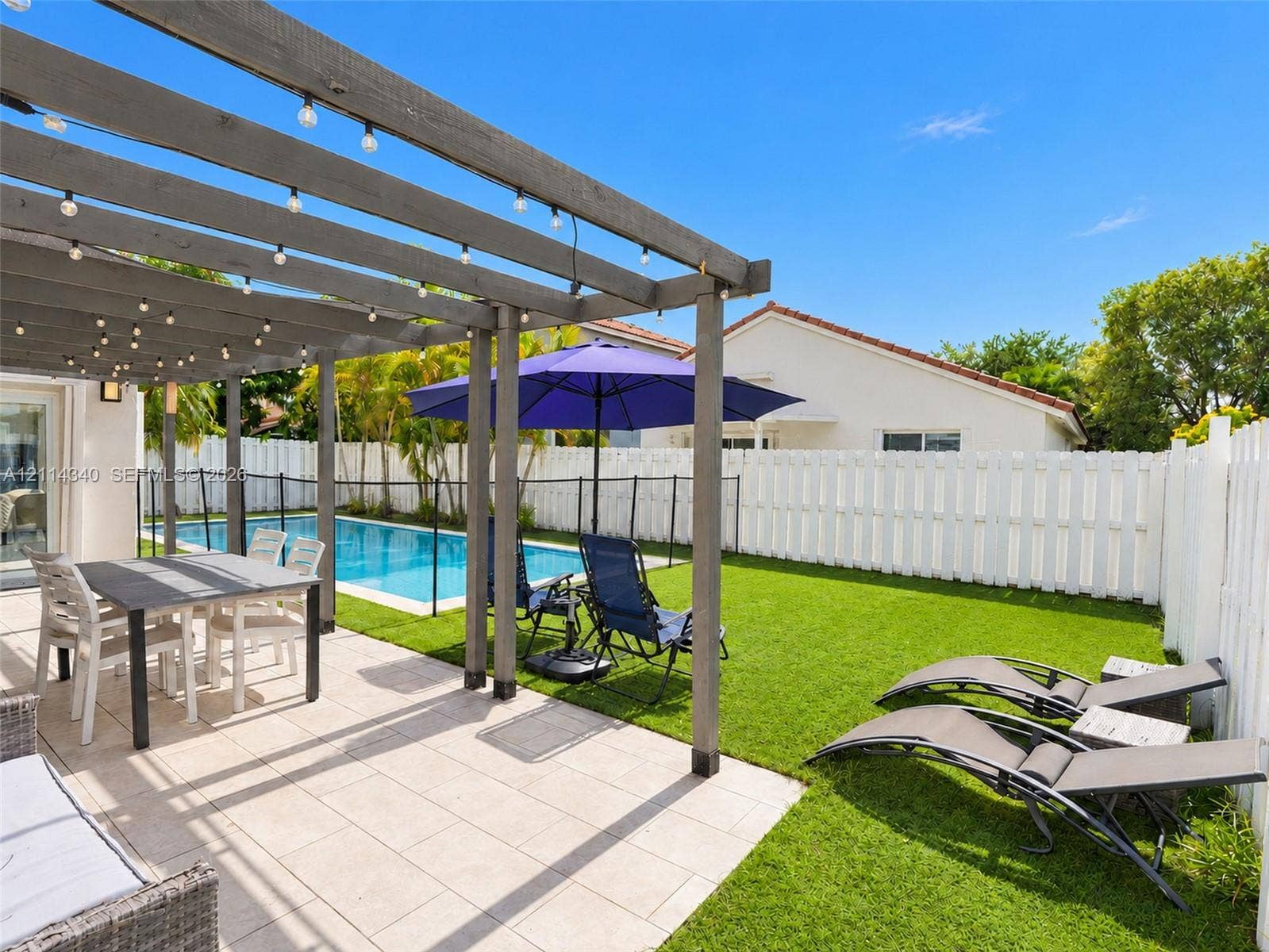 3586 SW 50th Street, Hollywood, FL 33312 Outdoor living space featuring a wood-beam pergola, light-toned stone patio, private swimming pool, synthetic lawn, and perimeter privacy fencing