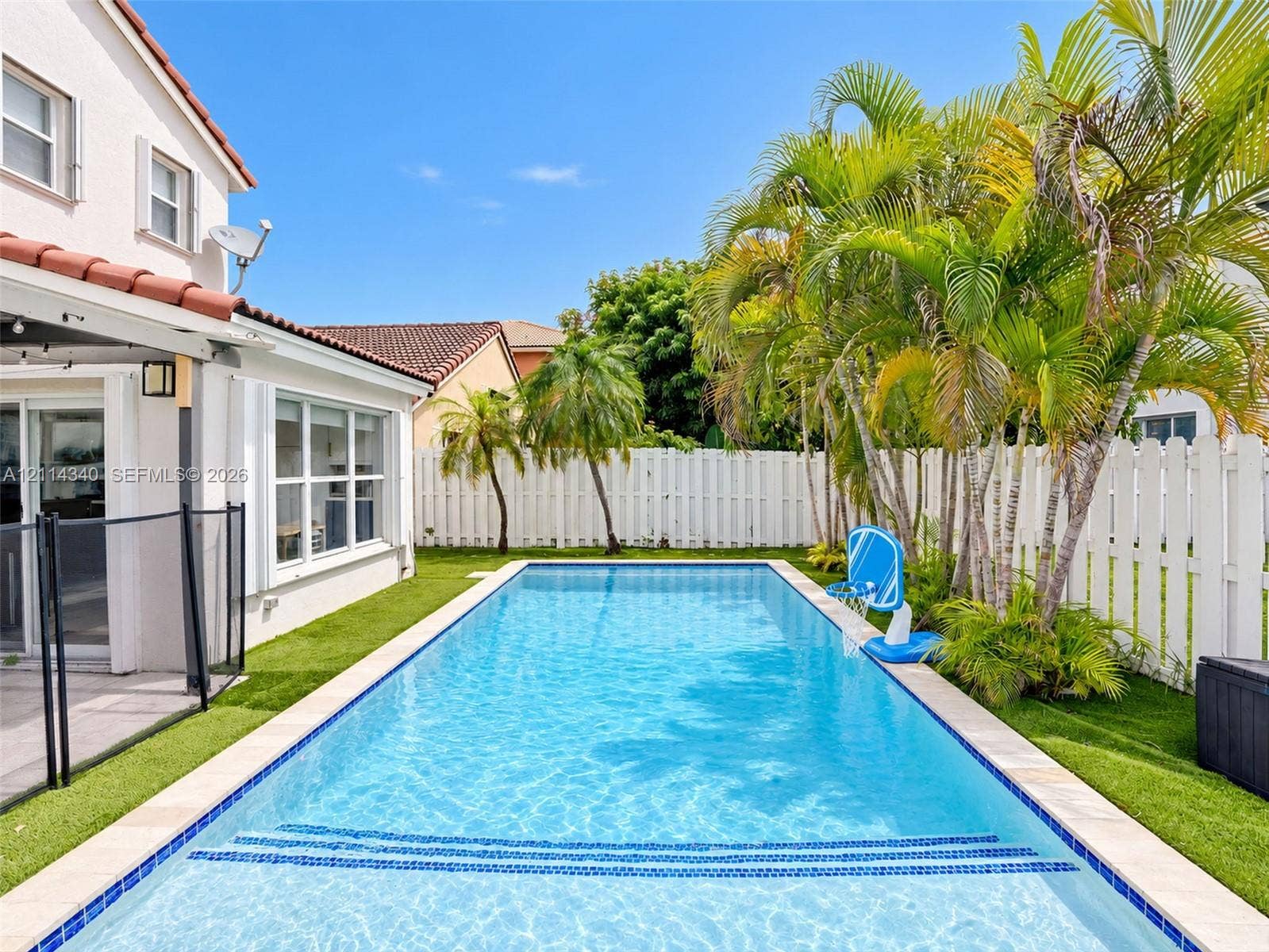 3586 SW 50th Street, Hollywood, FL 33312 Rectangular in-ground pool with stone coping and built-in steps, surrounded by artificial turf and mature palm trees, enclosed by a white privacy fence