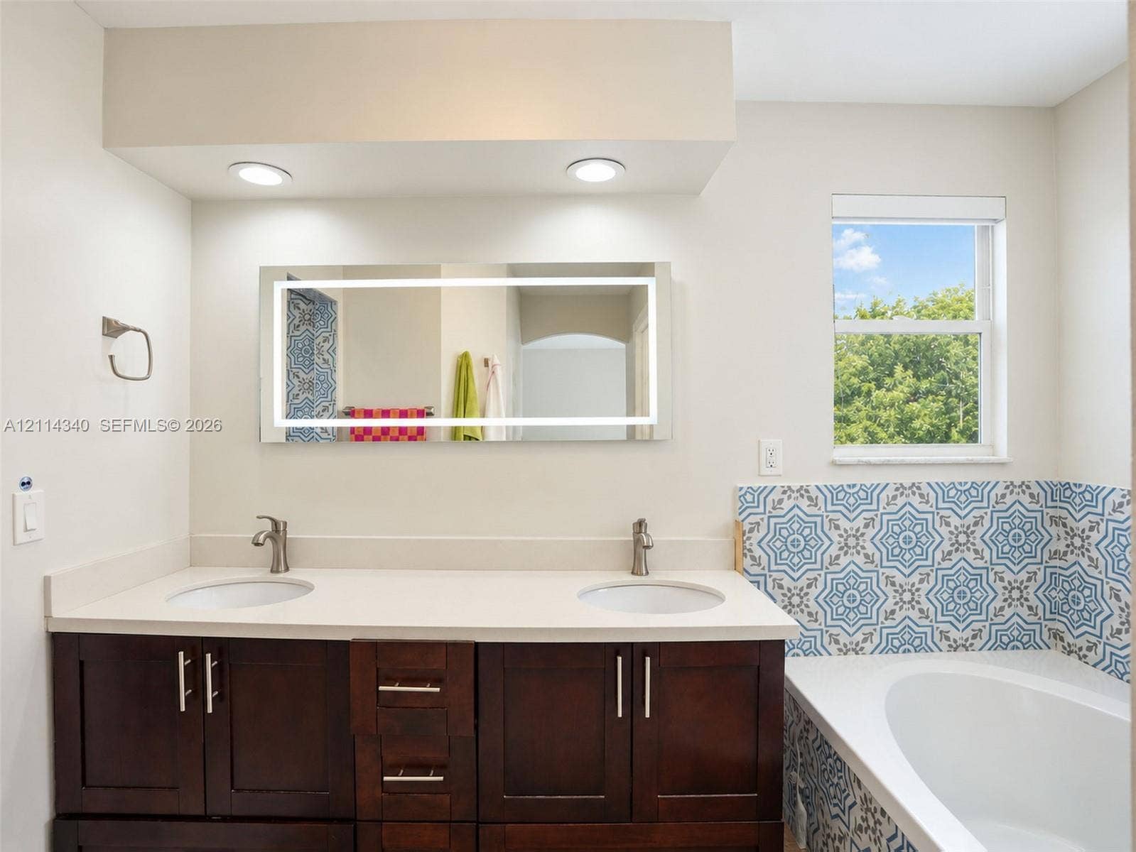 3586 SW 50th Street, Hollywood, FL 33312 Double vanity featuring a light-toned countertop, dual integrated sinks, dark wood-finish cabinetry, a backlit mirror, and decorative patterned tile surrounding the soaking tub