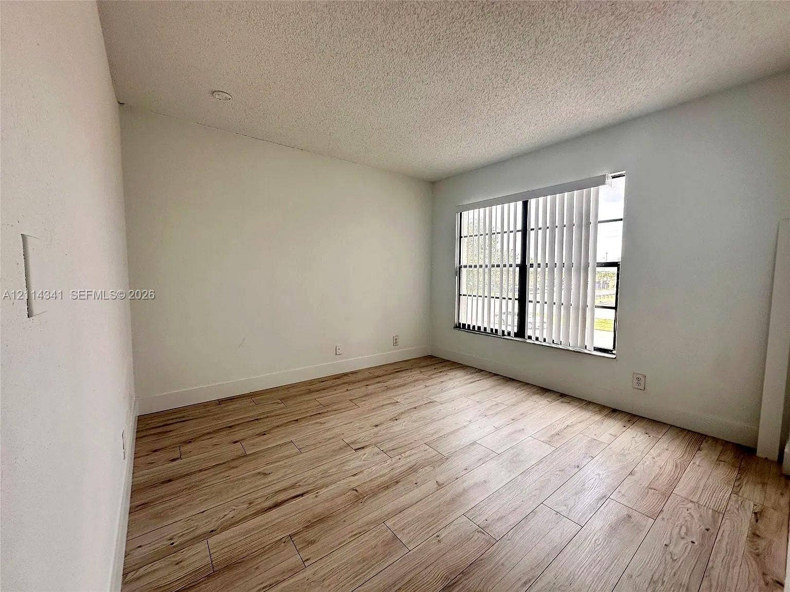 Room featuring wood-finish flooring, a multi-pane window with vertical blinds, and a textured ceiling