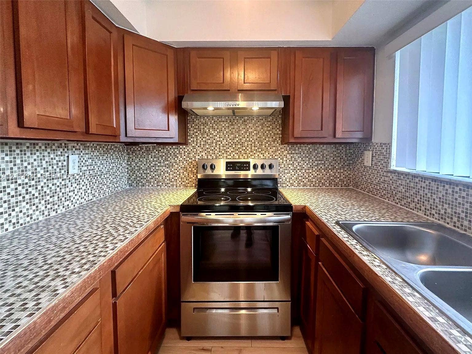 Kitchen featuring wood cabinetry, stainless steel electric range, stainless steel range hood, mosaic tile backsplash, and double-basin stainless steel sink