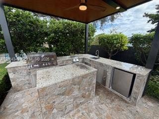 Outdoor kitchen under a covered structure featuring a natural stone-faced prep area, stone countertops, built-in stainless steel grill, undermount sink, and integrated refrigerator