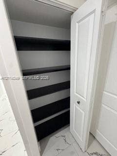 Storage closet featuring white paneled swing doors, multiple horizontal shelving tiers with dark-toned surfaces, and large-format tile flooring with veining detail