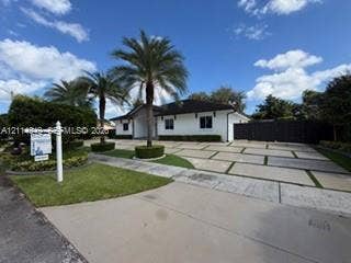 Single-story residence featuring a white exterior, dark shingle roof, expansive paver driveway with turf accents, and mature palm trees