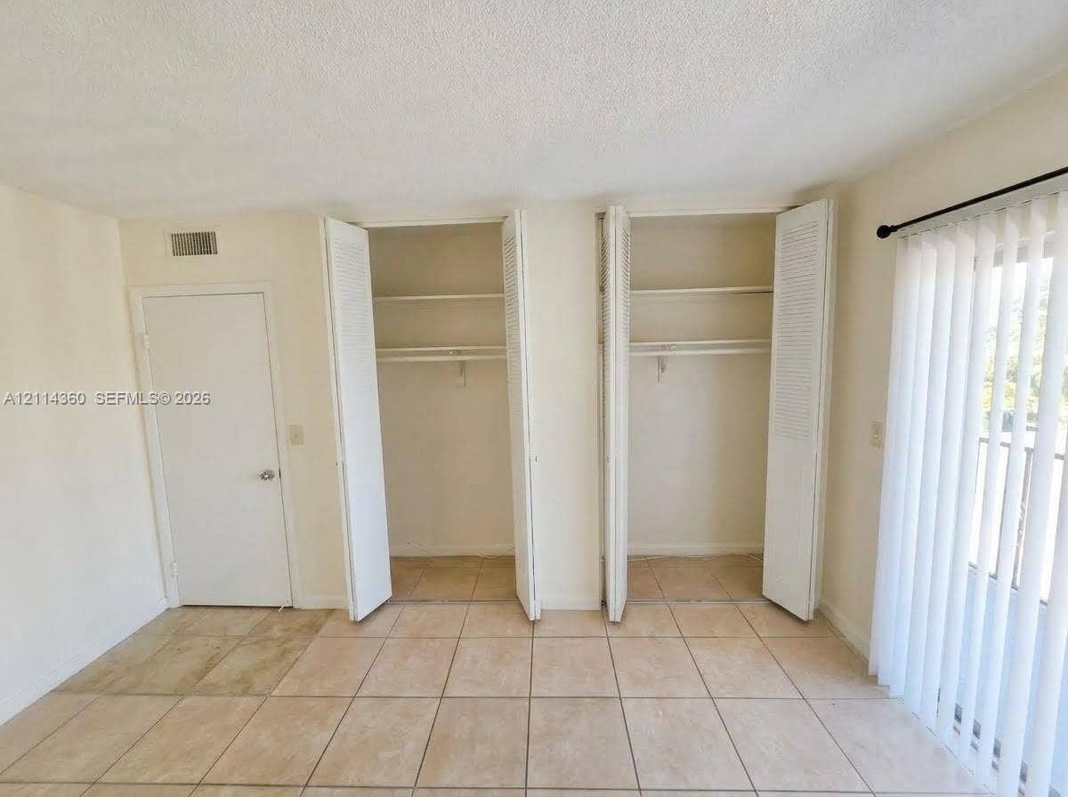 Dual closets with louvered bi-fold doors and interior shelving, tile flooring, and vertical blind window treatments