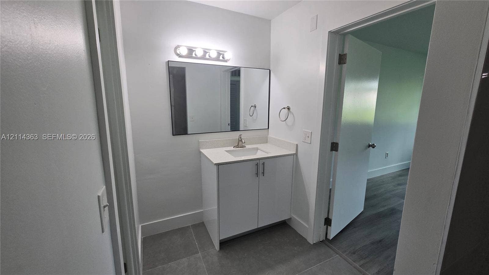 14840 Naranja Lakes Boulevard, #1G, Homestead, FL 33032 White vanity with stone-composite countertop, undermount sink, rectangular wall mirror, four-bulb vanity light fixture, and large-format tile flooring