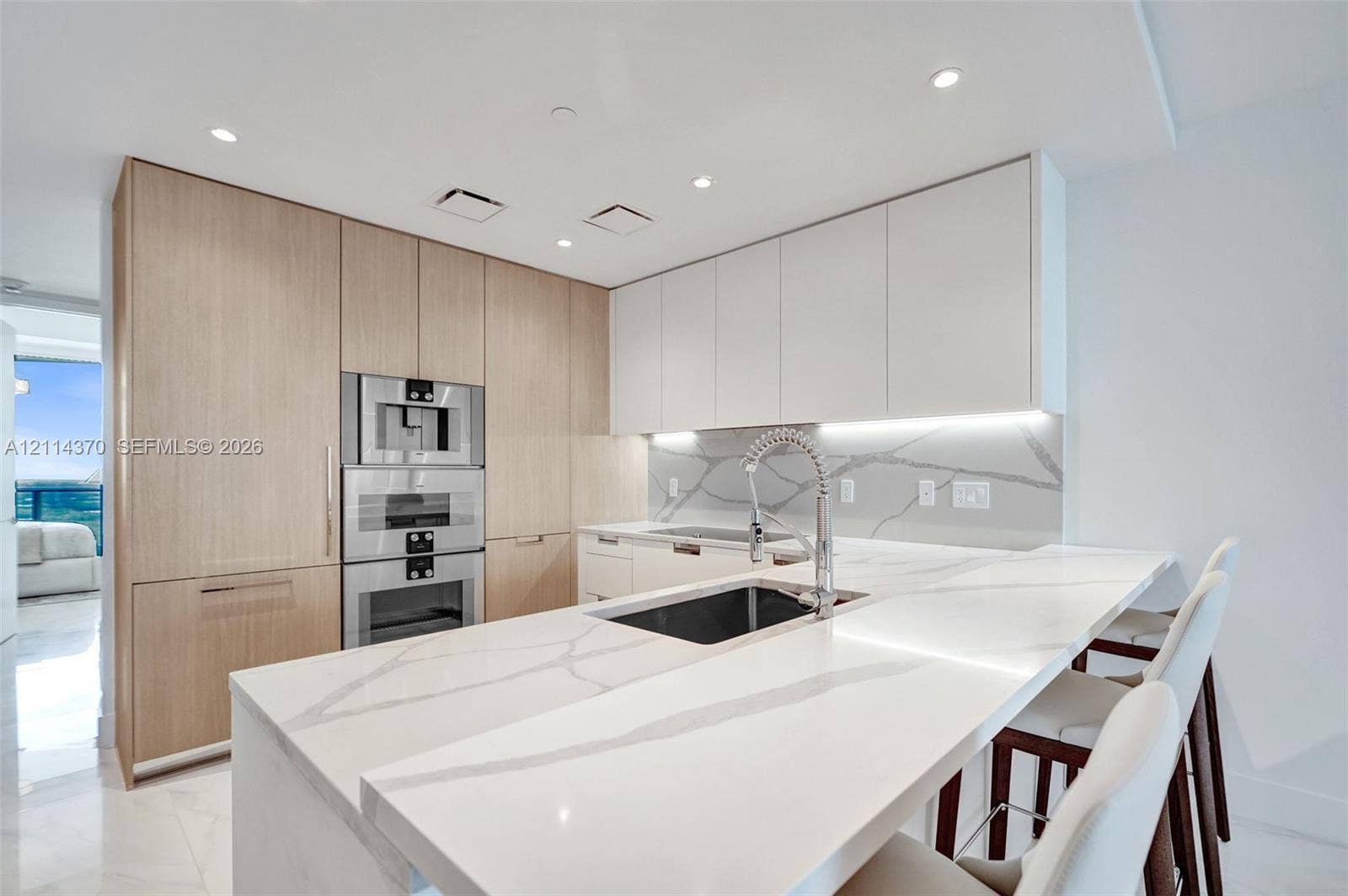 Sunny Isles Beach, Sunny Isles Beach, FL 33160 Kitchen featuring light-tone flat-panel cabinetry, white stone-finish waterfall island, integrated stainless steel double oven, and matching stone-finish backsplash with under-cabinet lighting