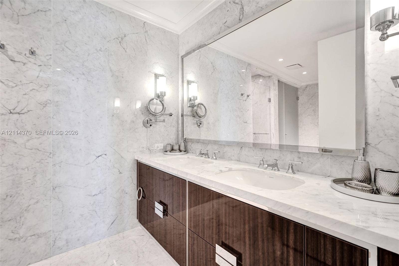 Sunny Isles Beach, Sunny Isles Beach, FL 33160 Dual-sink vanity with high-gloss cabinetry, stone-surfaced countertops, full-width wall mirror, wall-mounted lighting fixtures, and floor-to-ceiling stone wall tiling