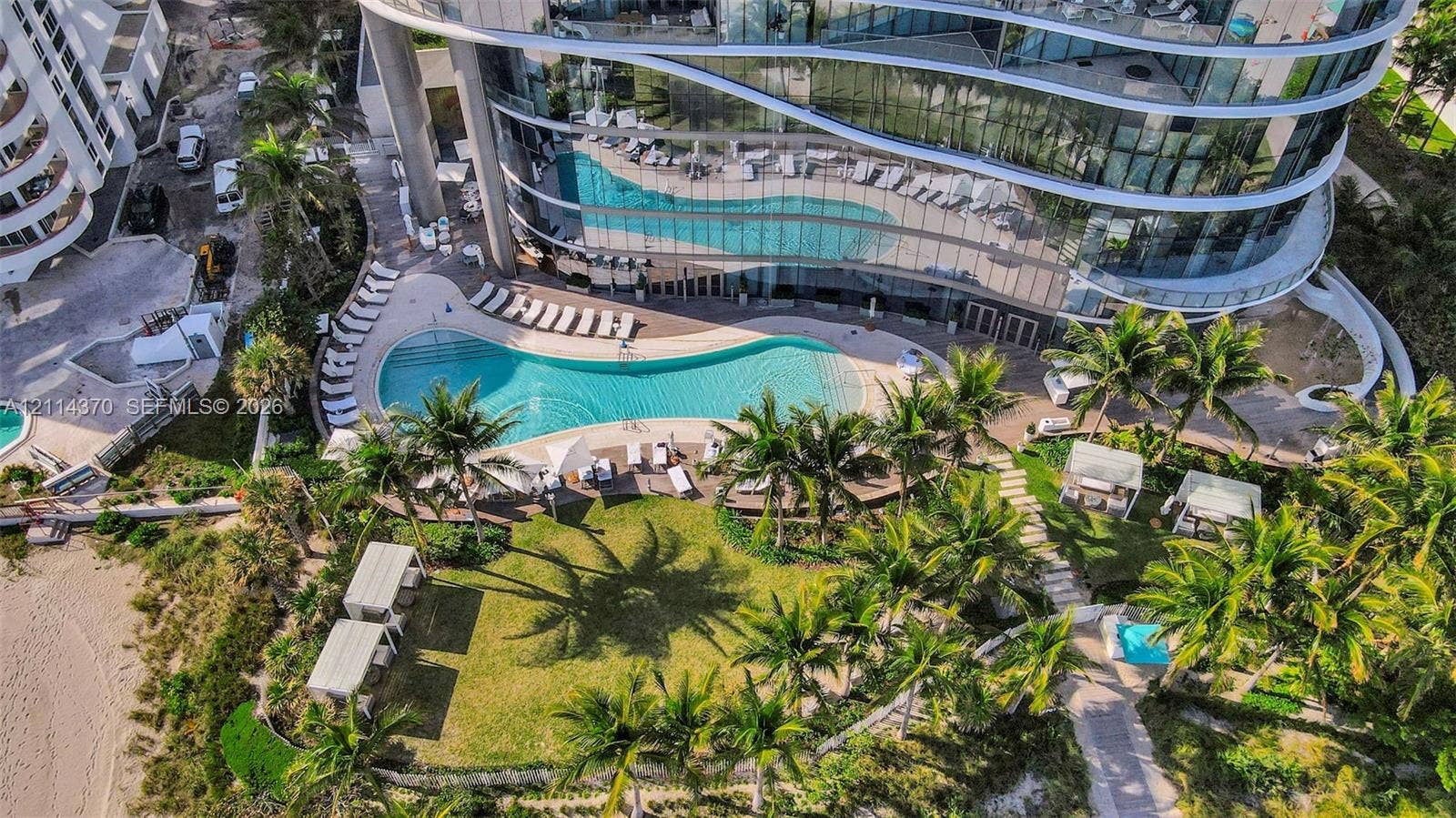 Sunny Isles Beach, Sunny Isles Beach, FL 33160 Curved high-rise building with floor-to-ceiling glass windows, expansive curved swimming pool, manicured lawn, palm-lined landscaping, and paved walkways